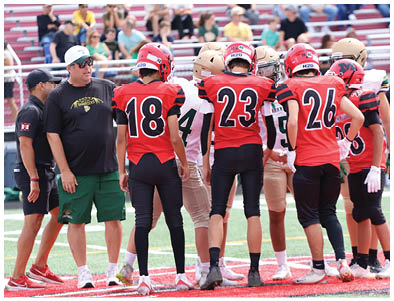 A group of people wearing football uniforms and helmets stand together on a field while two adults in casual sportswear talk nearby. Description generated by AI