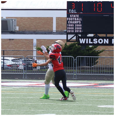 A person in sports gear is reaching out to catch a football near a goalpost on a field with a building and parked cars in the background. Description generated by AI