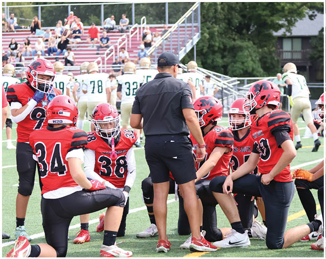A group of people wearing red and black football uniforms are kneeling and standing on a field while listening to a person in black clothing who appears to be coaching them. Description generated by AI