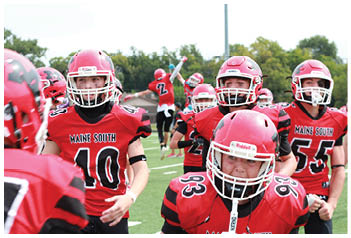 Several people wearing red and black football uniforms and helmets are gathered on a field with one person in the background jumping with an arm raised. Description generated by AI