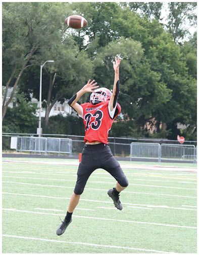 A person wearing a red and black football uniform with the number 23 is jumping with arms extended to catch a football on a grassy field with trees and buildings in the background. Description generated by AI