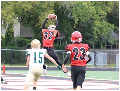 A person in a red and black football uniform is jumping to catch a football while three other players in white and green uniforms run toward them on a football field. Description generated by AI