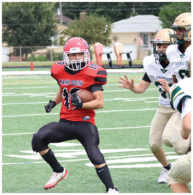 A person wearing a red and black football uniform with a helmet is running on a green football field while another person in a white and gold uniform reaches out toward them. Description generated by AI