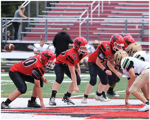 Children in red and white football uniforms are lined up on a field preparing for a play during a game. Description generated by AI