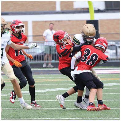 People in football uniforms are actively playing on a green field with one person running with the ball while others are preparing to block or tackle. Description generated by AI