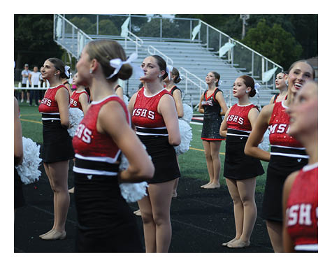 A group of adults in red and black cheerleading uniforms stand in formation on a track near a sports field with bleachers in the background. Description generated by AI