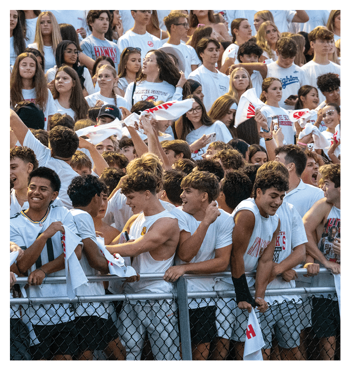 A large crowd of people wearing white shirts enthusiastically cheer and wave white towels behind a metal fence at an outdoor event. Description generated by AI