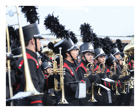 People in marching band uniforms with black hats and red accents are standing in a line holding brass instruments. Description generated by AI