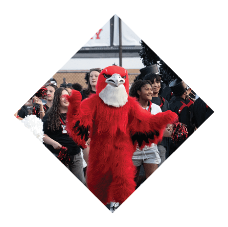 A person in a red bird mascot costume with yellow feet is standing with arms outstretched in front of a group of people, some holding pom-poms. Description generated by AI
