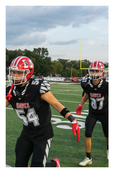 An adult wearing a black shirt, shorts, and a backpack is reaching out to shake hands with a person in a black football uniform and red helmet on a sports field. Description generated by AI