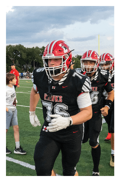 Adults wearing football uniforms with red helmets and black jerseys are walking on a field while another person in a red jersey and black shorts stands nearby. Description generated by AI