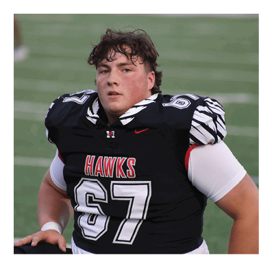 A person wearing a black football jersey with the number 67 and the word “HAWKS" stands on a field with a focused expression. Description generated by AI