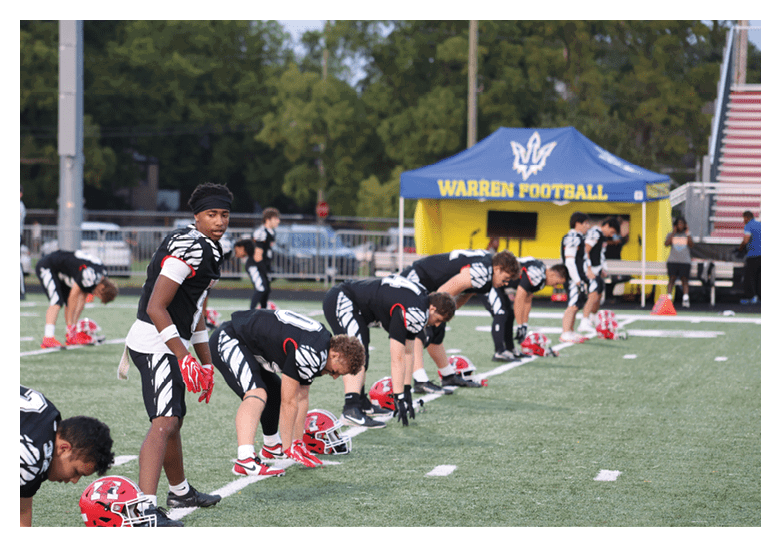 People in black and white football uniforms are lined up in a crouched position on a football field preparing for a play near a tent labeled Warren Football. Description generated by AI