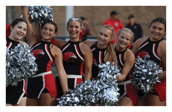 A group of six smiling people in matching cheerleading uniforms hold silver pom-poms and pose closely together outdoors. Description generated by AI