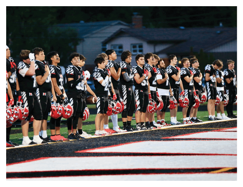 A group of people in black and white sports uniforms stand in a line on a football field holding red helmets, appearing to observe a moment of respect. Description generated by AI