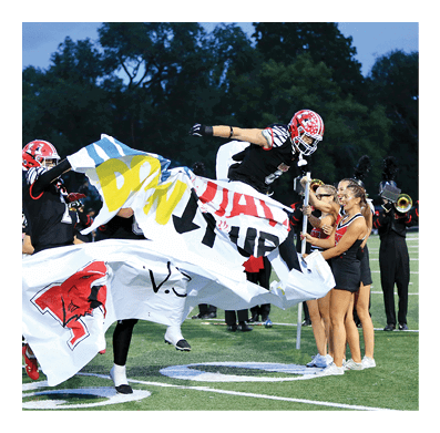 Two people in football uniforms jump through a torn banner on a field while cheerleaders and a marching band stand nearby. Description generated by AI