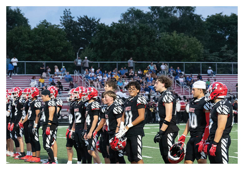 A group of people wearing black and red football uniforms stand in a line on a football field with spectators in the background. Description generated by AI