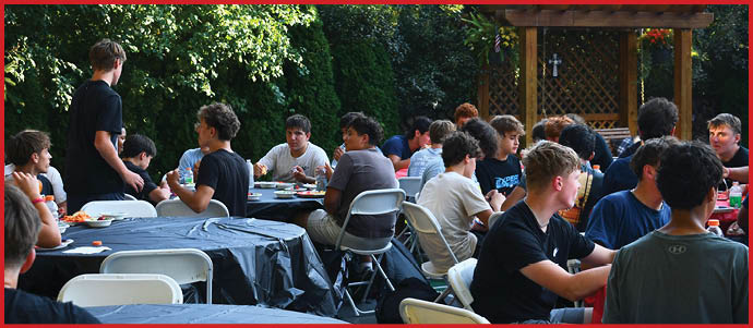A group of people sitting and standing around tables outdoors, engaged in conversation and eating in a shaded garden area. Description generated by AI