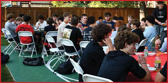 A group of people sitting on white folding chairs around tables outdoors in a backyard setting, engaged in conversation and eating. Description generated by AI