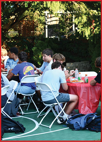 Several backpacks are placed on the ground near white folding chairs on a green outdoor sports court. Description generated by AI