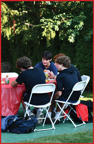 Three people with curly hair wearing dark clothing sit on white folding chairs around a red tablecloth-covered table outdoors, eating and talking, with backpacks on the ground nearby. Description generated by AI