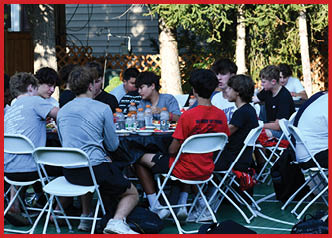A group of young people sit around a table outdoors, engaged in conversation and eating, with sports drinks and food containers on the table. Description generated by AI