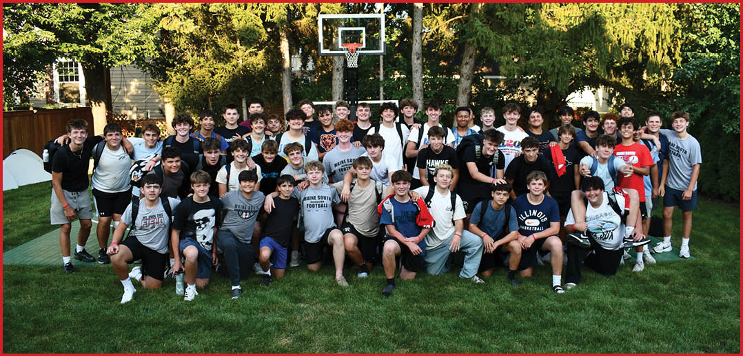 A group of people sitting around tables outdoors on a basketball court, eating and socializing under trees in a casual gathering. Description generated by AI
