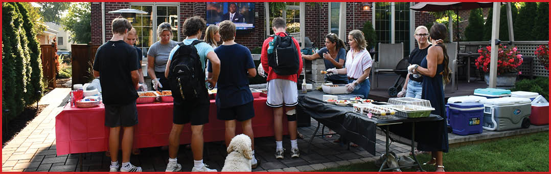 Several people stand outdoors in front of tables with food and coolers, while one person gestures as if speaking to the group. Description generated by AI