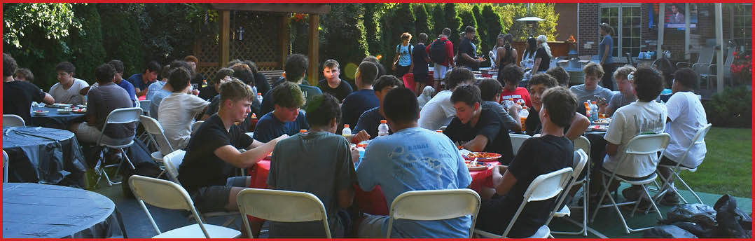 A large group of people sitting and eating together outdoors in a backyard setting with trees, a pergola, and a brick house in the background. Description generated by AI