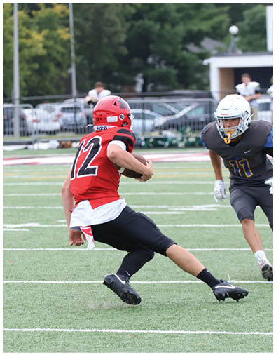A person in a dark sports uniform and white helmet is running on a football field while another person in a similar uniform is partially visible on the left side. Description generated by AI