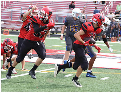 Two people wearing athletic shoes and sportswear are running or playing on a green turf field with white and red markings. Description generated by AI