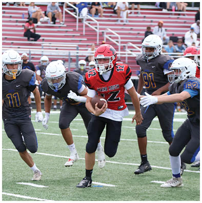 People wearing sports uniforms are running on a football field with one person holding a football. Description generated by AI
