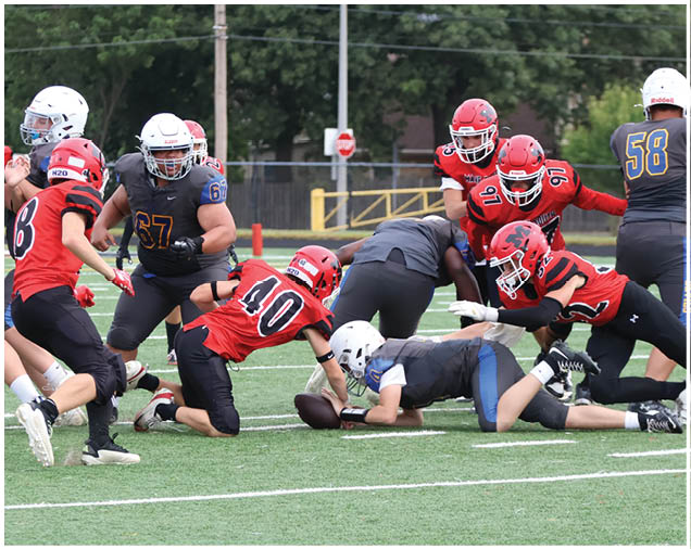Several people wearing helmets and sports uniforms are engaged in a football game on a green field, with some players tackling others near the ground. Description generated by AI
