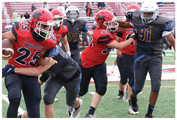 Players in red and black uniforms and players in dark gray uniforms are actively competing in a football game on a green field with white yard lines. Description generated by AI