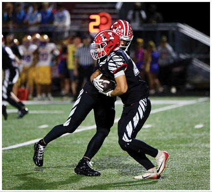 Two people wearing black and white football uniforms with red helmets are engaged in a play on a football field at night. Description generated by AI