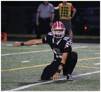 A person in a black football uniform with a red and white helmet is crouched on a football field holding a football in a kicking position with a referee and another person in the background near a down marker showing the number four. Description generated by AI