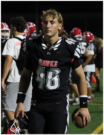 A person wearing a black football uniform with the number 48 and the word “HAWKS" on the front stands on a field with other players in the background. Description generated by AI