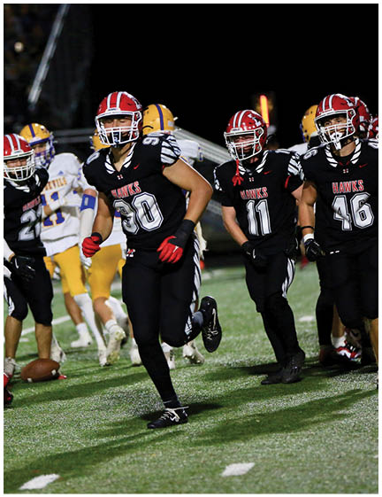 Three people in black football uniforms with red helmets and gloves are on a field with two people in white and yellow uniforms in the background near a football on the ground. Description generated by AI