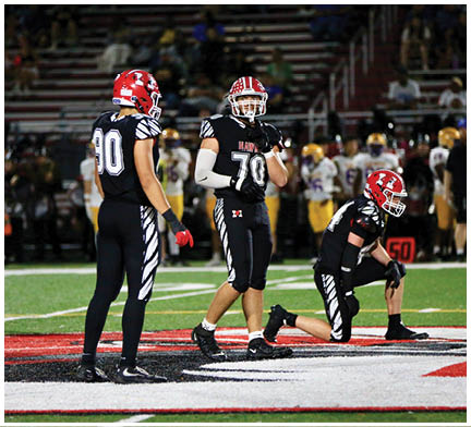 Three people in black football uniforms with white and zebra-striped accents stand and kneel on a football field during a game. Description generated by AI