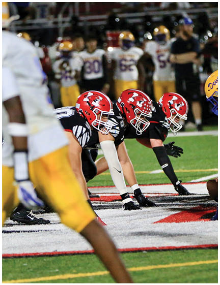 Three football players in red helmets and black uniforms are lined up on the field facing an opposing player in a white jersey and yellow pants during a game. Description generated by AI