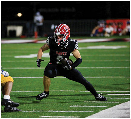 A person wearing a red and white helmet and black sports uniform with the word “HAWKS" is running on a football field while holding a football. Description generated by AI