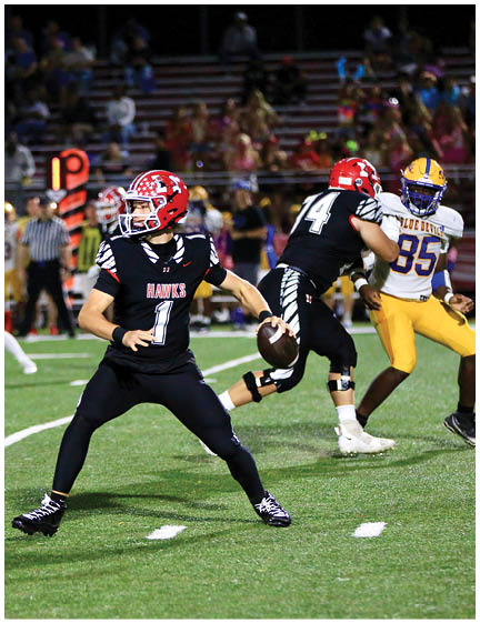 A person wearing a black football uniform with a red helmet is preparing to throw a football on a field while another player in a similar uniform blocks an opponent in yellow and purple. Description generated by AI