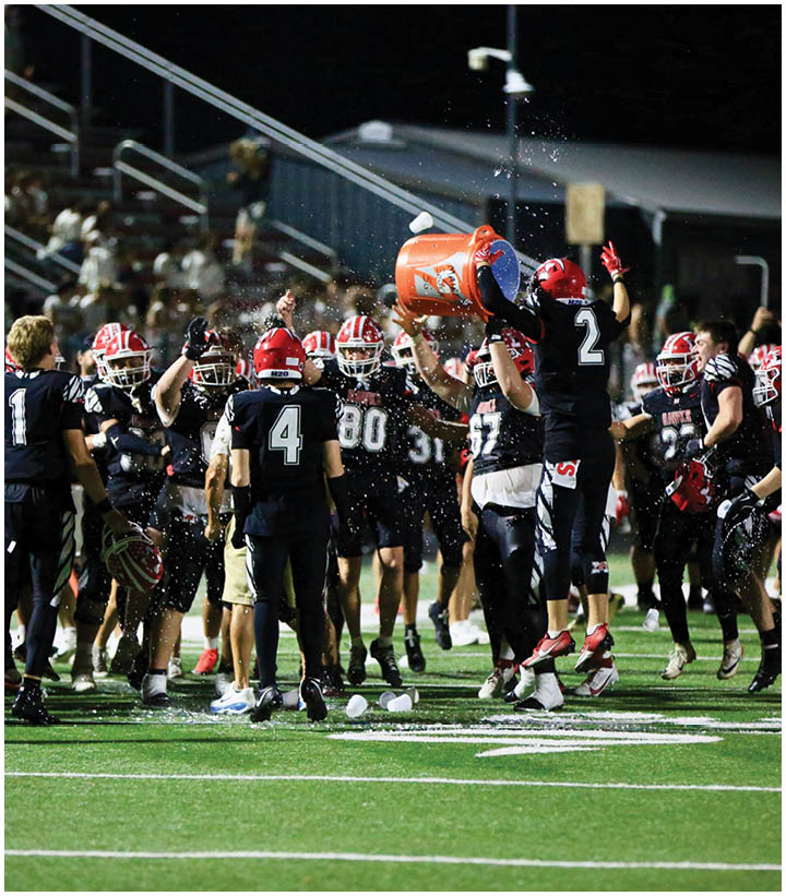 A group of people wearing football uniforms and helmets are gathered on a field at night while one person pours water from a large cooler over another. Description generated by AI