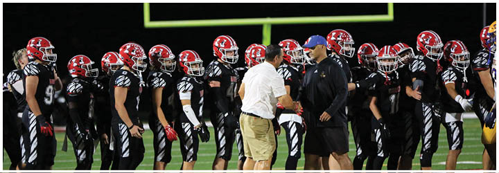 A group of people wearing black and red football uniforms stand in a line on a field while two adults in casual clothing shake hands near the center. Description generated by AI