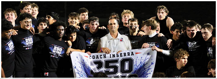 A group of people wearing matching black shirts surround an adult holding a banner that reads “Coach Inserra 250 Wins" in a celebratory setting. Description generated by AI