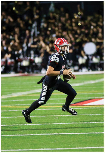 A person wearing a red helmet and black football uniform is running on a football field while holding a football. Description generated by AI