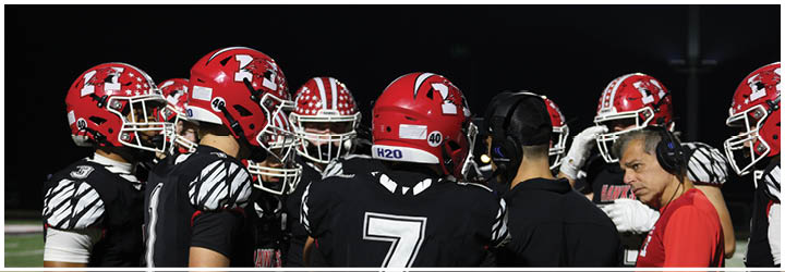 A group of people wearing red helmets and black football uniforms are gathered closely on a field at night, seemingly in a team huddle or discussion. Description generated by AI