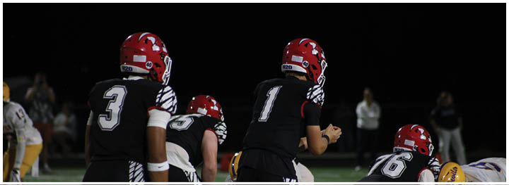 Several people in black sports uniforms with white stripes are positioned on a football field facing opponents in yellow and white uniforms during a game. Description generated by AI