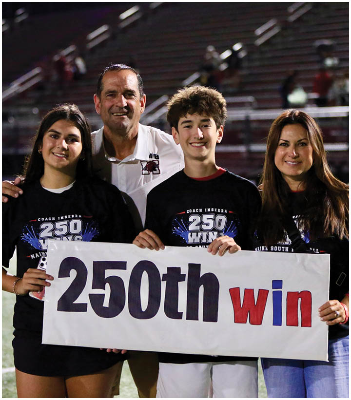 Four people smiling on a sports field at night hold a sign celebrating a milestone win. Description generated by AI