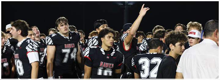 A group of people wearing black football uniforms with “HAWKS" written on them are gathered together on a field at night. Description generated by AI